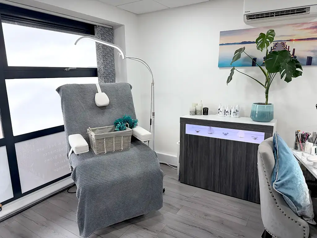 A modern beauty treatment room with a reclining chair and grey cover awaits, complete with a cupboard of products, scenic wall art, and a calming potted plant—the perfect space for your trusted beauty therapist in Ipswich.