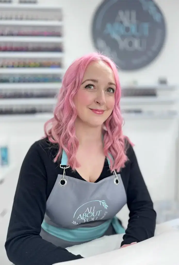 A woman with pink hair, wearing a black shirt and a grey "All About You" apron, sits at a table in a bright beauty therapist Ipswich salon, smiling at the camera. Shelves of nail varnish are visible in the background.