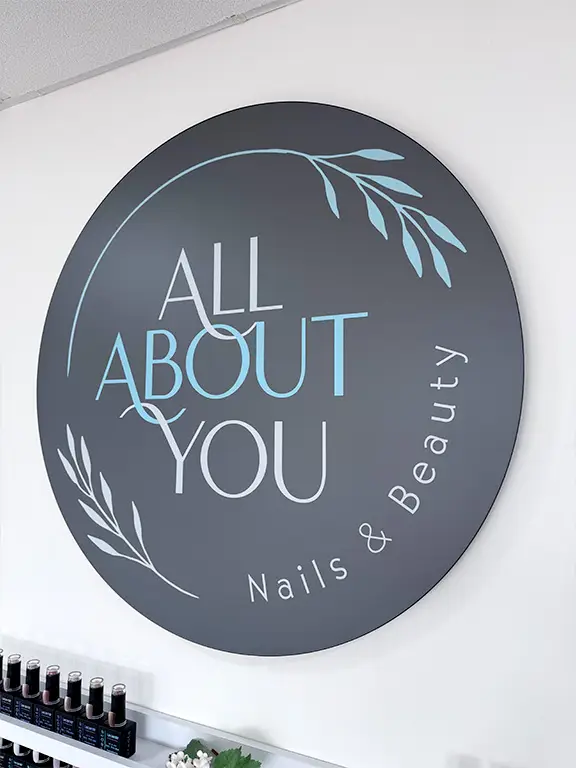A large round sign on a white wall reads ALL ABOUT YOU Nails & Beauty in white and light blue text, decorated with simple leaf graphics. Small bottles are visible on a shelf below the sign.