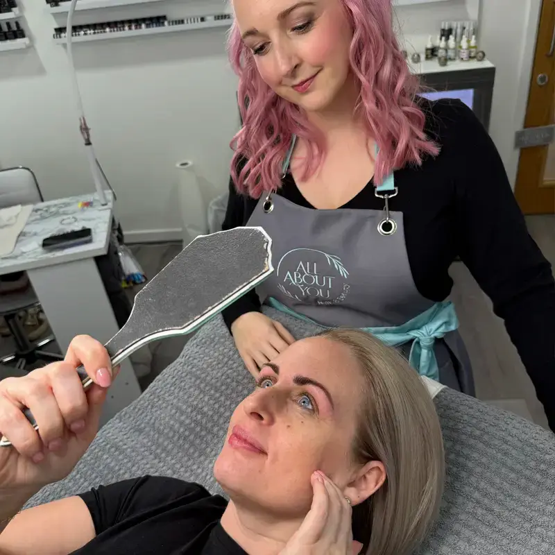 A woman with pink hair in a salon apron stands behind a client holding a handheld mirror, as the client examines her freshly done eyebrows and smiles whilst lying on a treatment bed.