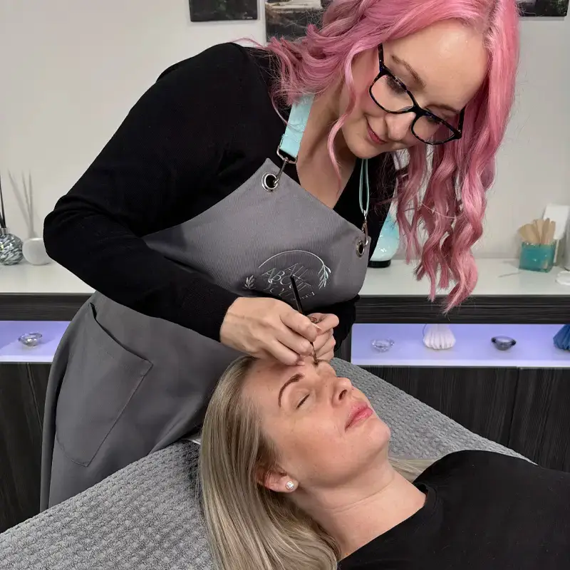 A woman with pink hair and glasses, wearing a grey apron, carefully shapes the eyebrows of a blonde woman lying on a treatment bed in a beauty salon.