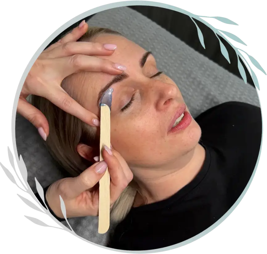 A woman with closed eyes lies on a grey surface while someone applies wax to her eyebrow with a wooden spatula, preparing for an eyebrow waxing treatment at Hands & Feet Treatments Ipswich.
