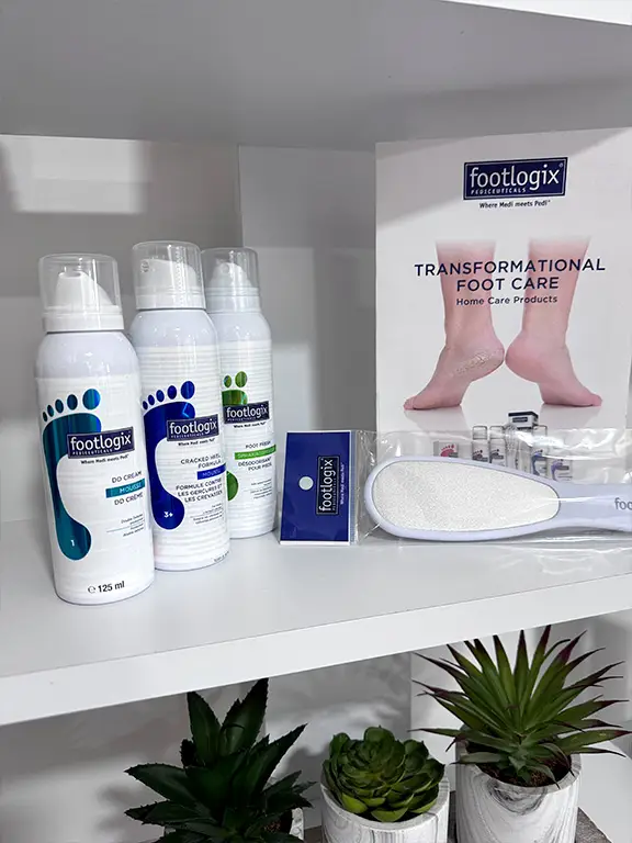 Foot care products, including three Footlogix bottles, a foot file, a brochure titled Transformational Foot Care, and small potted plants, arranged on white shelves.