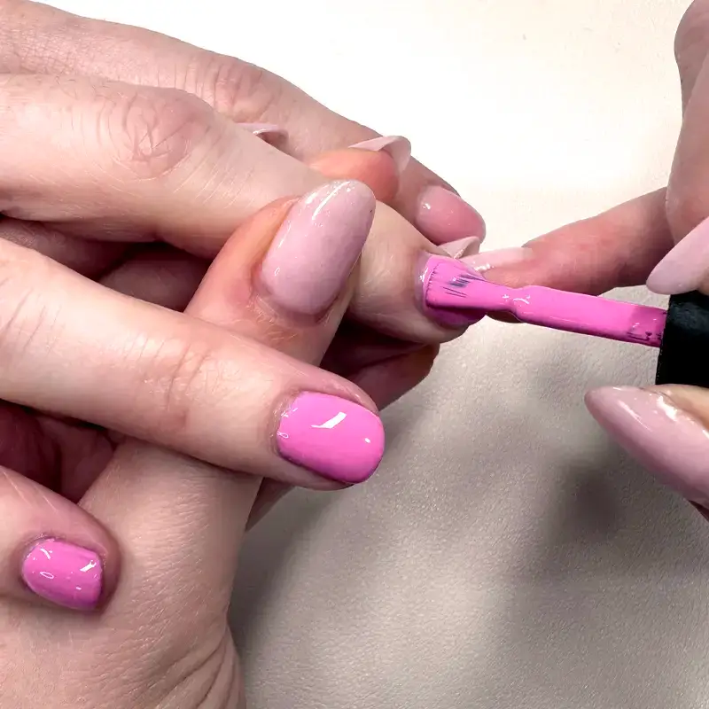 A close-up of a person painting another person's fingernails with bright pink nail varnish, while holding their hand steady. The nails being painted are already shaped and some are painted.
