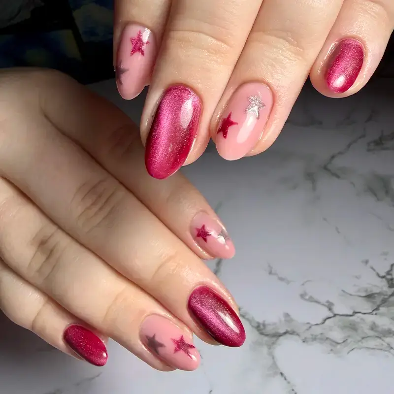 A hand with pink and deep magenta almond-shaped nails, some featuring shiny finishes and others decorated with metallic star stickers, displayed on a marble surface.