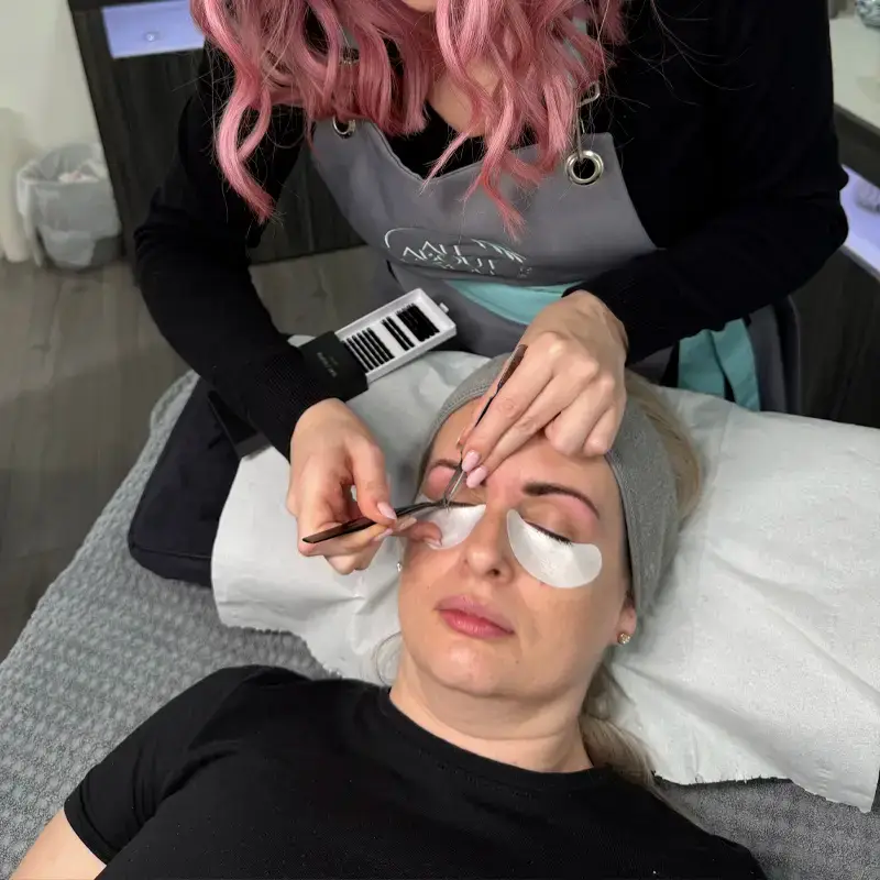 A woman with pink hair applies eyelash extensions to a client lying on a salon bed. The client has eye pads under her eyes and is resting with her eyes closed.