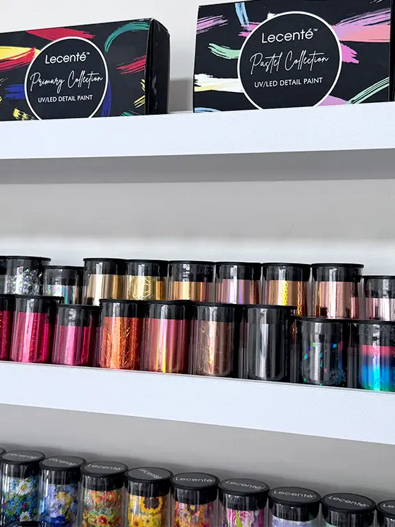 Shelves display colourful jars of nail art foils and glitter. Two boxes labelled Lecenté Primary Collection and Lecenté Pastel Collection UV/LED Detail Paint sit on the top shelf.