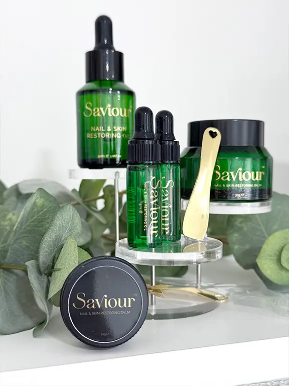 Four green Saviour skincare products with gold accents are displayed on a clear stand, surrounded by eucalyptus leaves. Included are dropper bottles, a jar, a balm tin, and a gold applicator spatula with a heart cut-out.