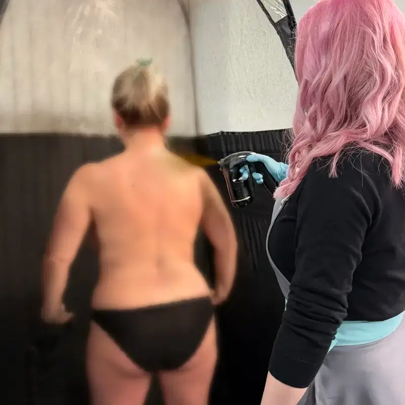 A person with pink hair applies a spray tan to the back of a person wearing black bikini bottoms, standing in a spray tan booth.